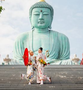Two Asian young woman with japanese style dress stay on stair in front of green big buddha statue with day light and they look happy.